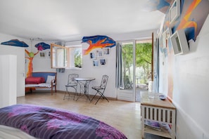 Et værelse med en seng, en sofa, et spisebord med stole og et stort vindue med udsigt over grøn vegetation.