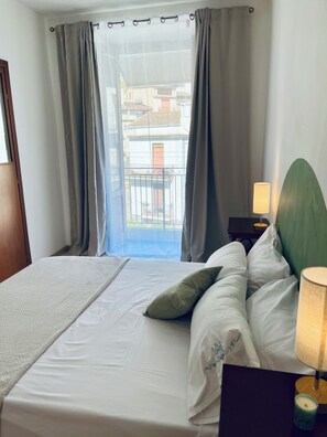 A hotel room with a bed, pillows, a bedside lamp, and a view of buildings through the window. - La Fontanella - Authentic Sicilan Retreat (Ciminna)