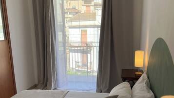 Camera d'albergo con un letto, una lampada da comodino, un comodino e vista sui palazzi attraverso tende trasparenti.