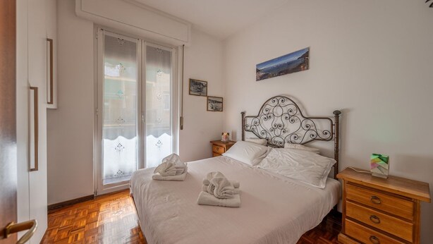 Quarto com cama, piso de madeira, mesa de cabeceira, cômoda e janela com vidro fosco.