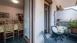 Varanda com mesa e cadeiras, um vaso de planta e uma porta de vidro deslizante que dá para um espaço interno com mesa e cadeiras de jantar.