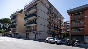 Um edifício residencial com vários andares, varandas, uma rua em frente e carros estacionados.