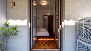 Corredor estreito que dá para um quarto, com uma cama, um vaso de plantas e um ponto de luz na parede.
