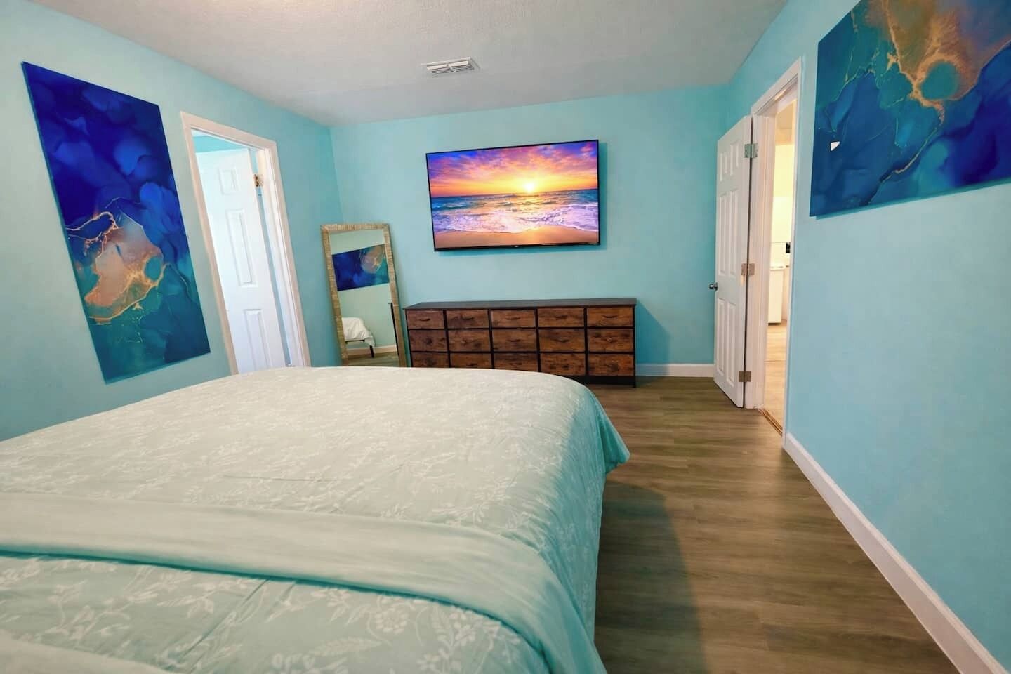 A bedroom with a bed, a dresser, a painting of a sunset, and a door.