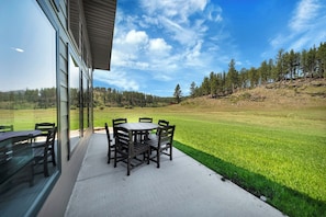 Um pátio com uma mesa redonda e cadeiras, uma mesa de vidro e vista para um campo gramado e árvores.