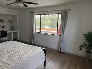 Ein Schlafzimmer mit Bett, Deckenventilator, Fenster mit Vorhängen, Schreibtisch mit Stuhl und einer Zimmerpflanze.