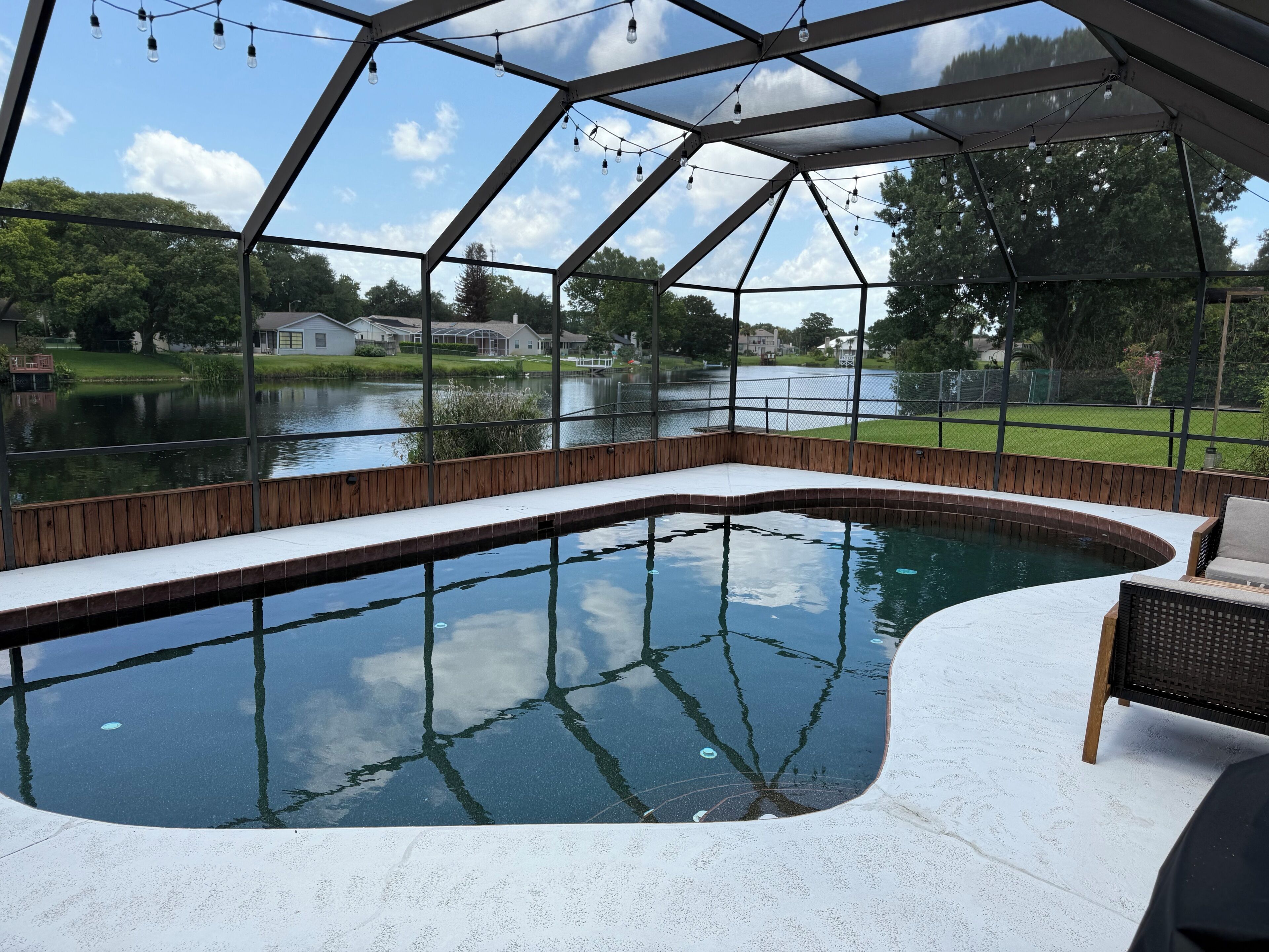 A swimming pool with a wooden deck and a glass enclosure, surrounded by a fence and a view of a lake and houses.