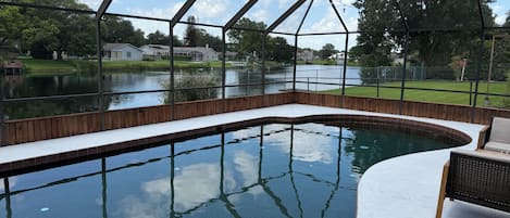 A swimming pool with a wooden deck and a glass enclosure, surrounded by a fence and a view of a lake and houses.