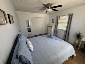 Ein Schlafzimmer mit Bett, Deckenventilator, Kleiderschrank, Schminktisch und einem Fenster mit Jalousien.