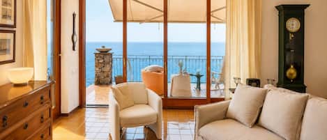 A living room with a sofa, armchairs, a wooden dresser, and a view of the sea through a glass door.