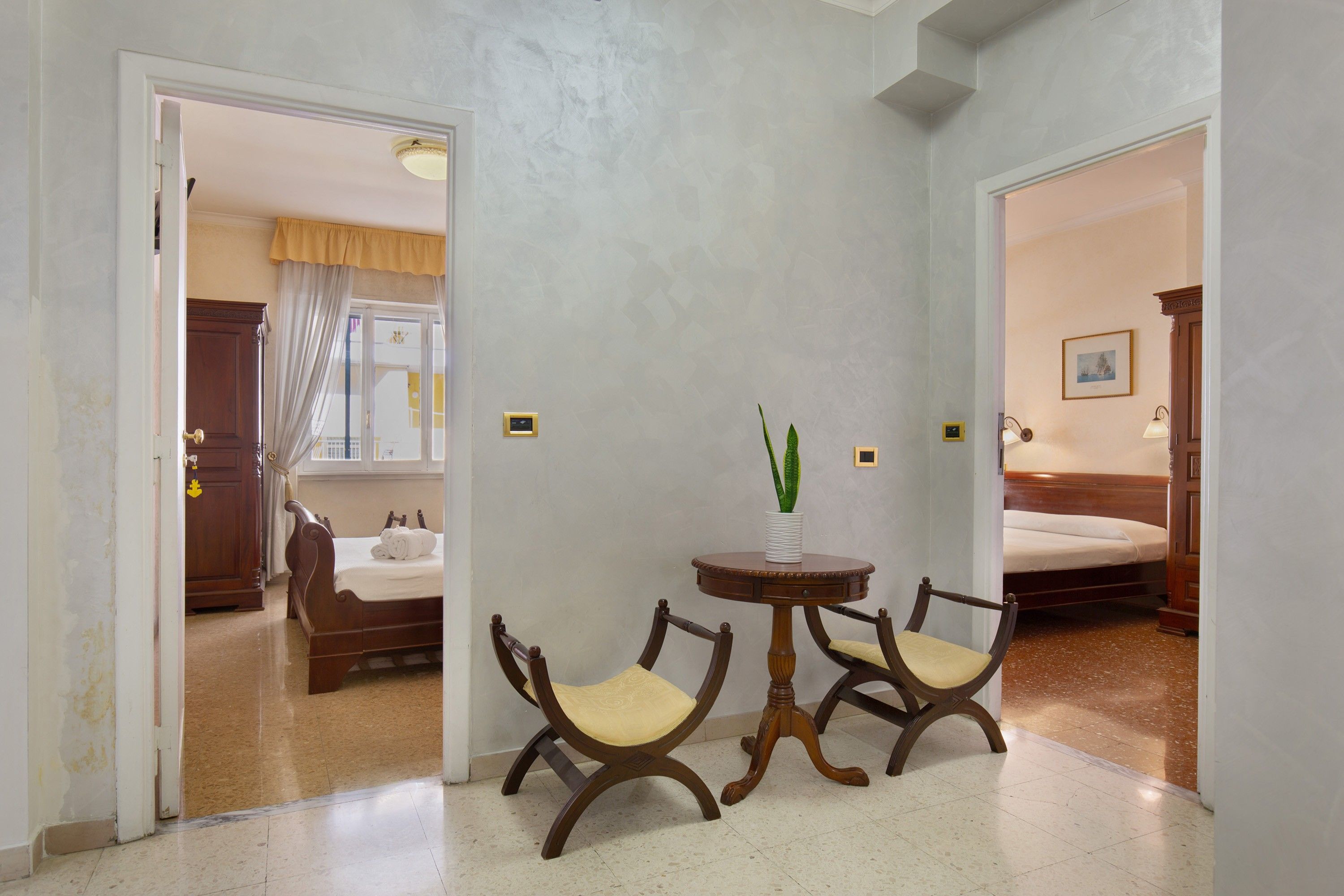 A hotel room with a bed, a bedside table with a vase, two chairs, and a door leading to another room.