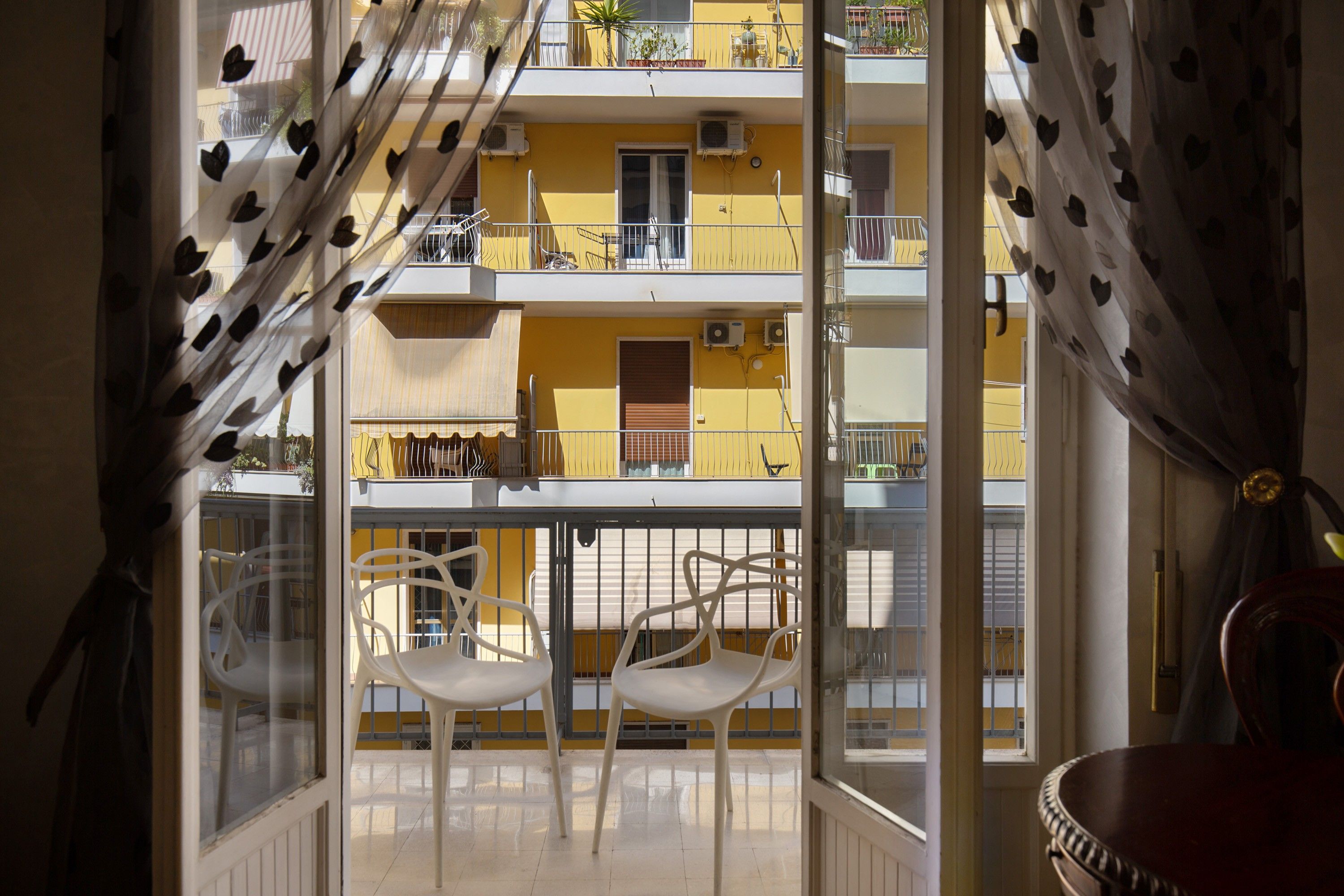 A balcony with two white chairs, a yellow building in the background, and a view through a window with curtains.