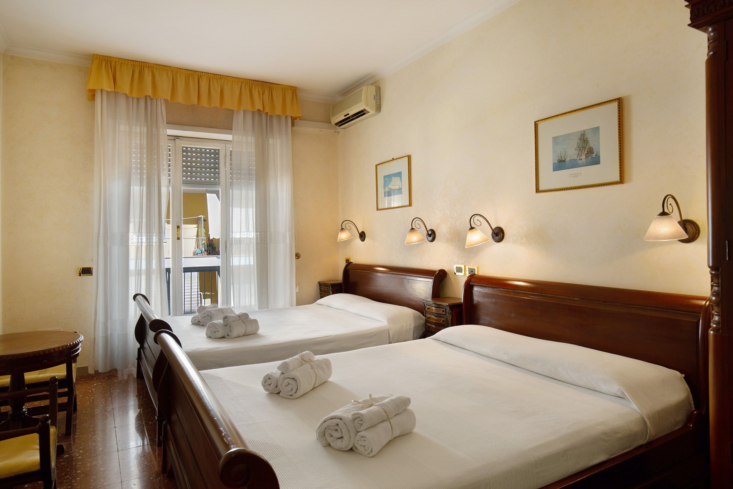 Ein Hotelzimmer mit zwei Betten, einem Holzkopfende, einem Tisch und einem Stuhl. Auf den Betten liegen Handtücher und es gibt ein Fenster mit Vorhängen.