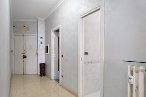Ein Flur mit weißen Wänden, einem Marmorboden und Türen zu Zimmern. An der Wand befinden sich Lichtschalter und eine Heizung.