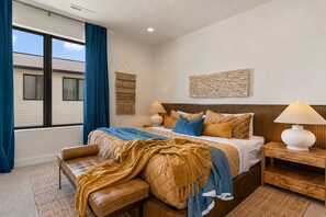 A bedroom with a large bed, two bedside tables, a bench, a window with curtains, and a wall-mounted lamp. - Red Rock Isle: Private Pool + Hot Tub! Games! (Washington)