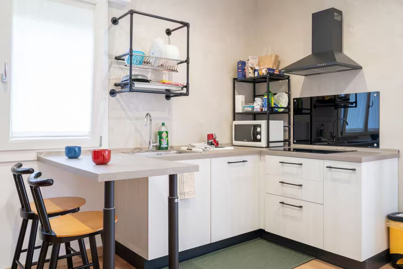Una cocina compacta con muebles blancos, una campana extractora negra, un microondas y un escurreplatos.