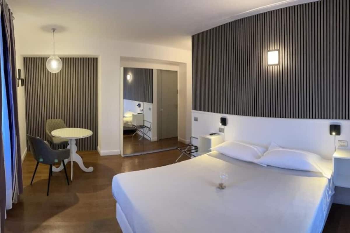A hotel room with a large bed, bedside tables, a desk with a chair, and a mirror.