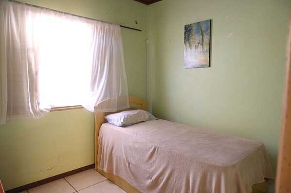 A small, single-bed room with a plain bed, a window with sheer curtains, and a painting on the wall.