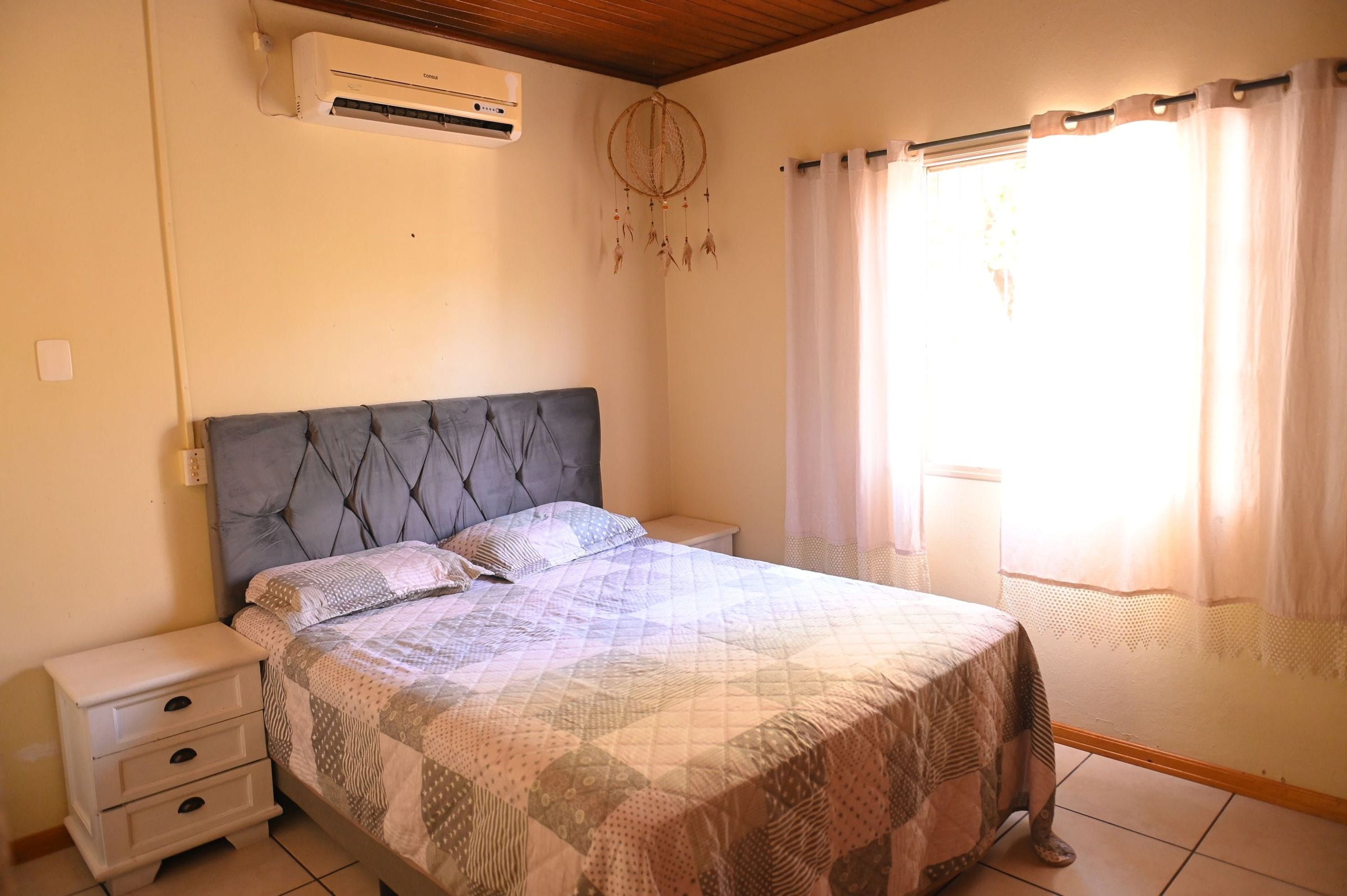 A bedroom with a bed, bedside tables, an air conditioner, a dreamcatcher, and a window with curtains.