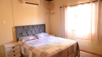 A bedroom with a bed, bedside tables, an air conditioner, a dreamcatcher, and a window with curtains.