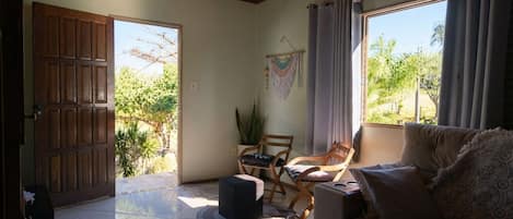 A room with a wooden door, a sofa, two chairs, a small table, and a view of greenery outside.