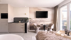 Een moderne hotelkamer met een keuken, een grote flatscreen televisie, een zithoek met twee stoelen, een groot raam en een bad.