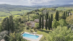 Ein Pool, umgeben von einem Garten mit hohen Bäumen, ein Steinhaus und Blick auf sanfte Hügel.
