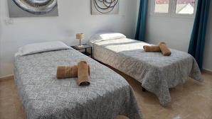 A room with two beds, each with a patterned bedspread and a rolled towel. There are abstract paintings on the walls, a small table with a lamp, and a window with curtains.