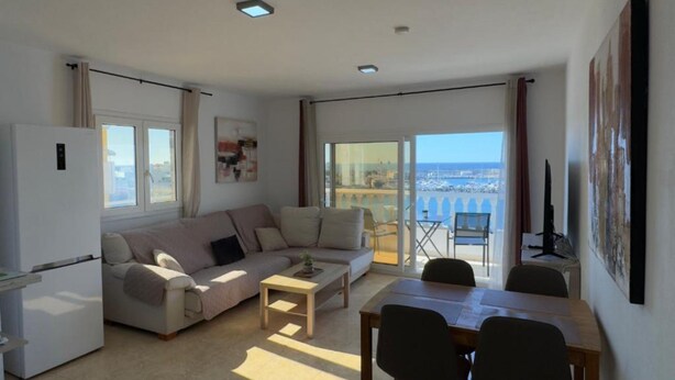 Una sala de estar moderna con sofá, mesa de centro y comedor. Hay un balcón con vistas al mar.