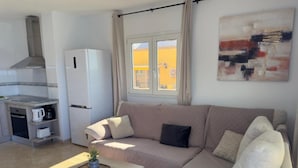 A living room with a sofa, a coffee table, a kitchen area with a refrigerator, and a window with curtains.