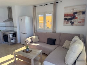 A living room with a sofa, a coffee table, a kitchen area with a refrigerator, and a window with curtains.