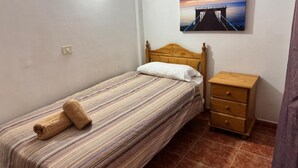 Una cama individual con una colcha a rayas, una mesita de noche de madera con cajones, una lámpara de pared y un cuadro de un muelle al atardecer.