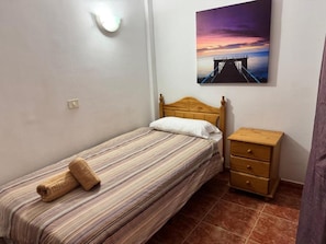 Een eenpersoonsbed met een gestreepte spreidek, een nachtkastje van hout met lades, een wandlamp en een schilderij van een pier bij zonsondergang.