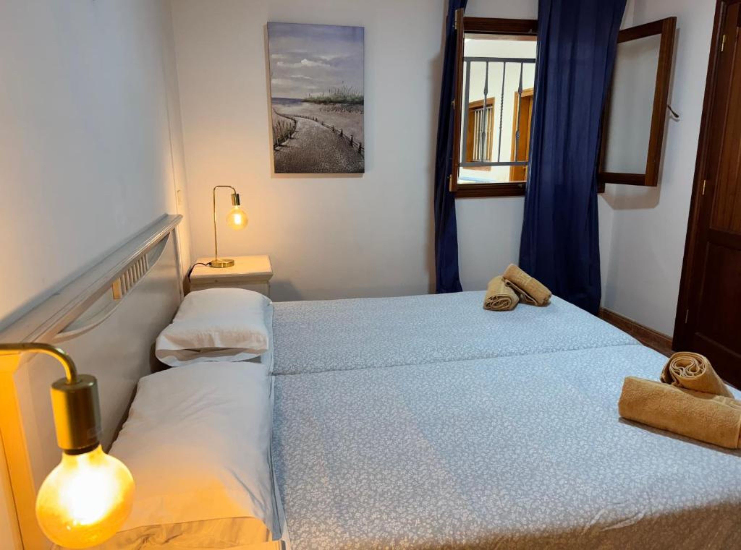 Une chambre d’hôtel avec un lit, une table de chevet, une lampe, un tableau au mur et une fenêtre avec des rideaux.