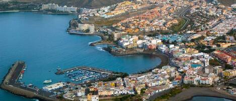 An aerial view of a coastal town with a marina, buildings, and a hilly backdrop.