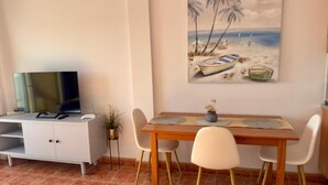 Un comedor con una mesa de madera, cuatro sillas, un cuadro de una escena de playa y un televisor sobre un mueble.