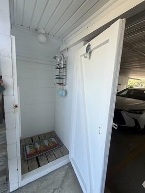 Une porte blanche avec un paillasson, un petit espace douche avec un pommeau de douche et un porte-serviettes, et une voiture garée à l’intérieur d’un garage.