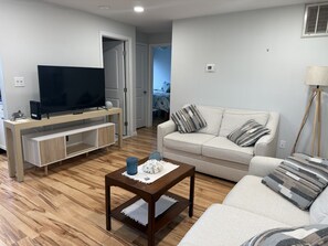 Una sala de estar con un televisor de pantalla plana sobre un soporte de madera, un sofá blanco con cojines a rayas y una mesa de centro con adornos.