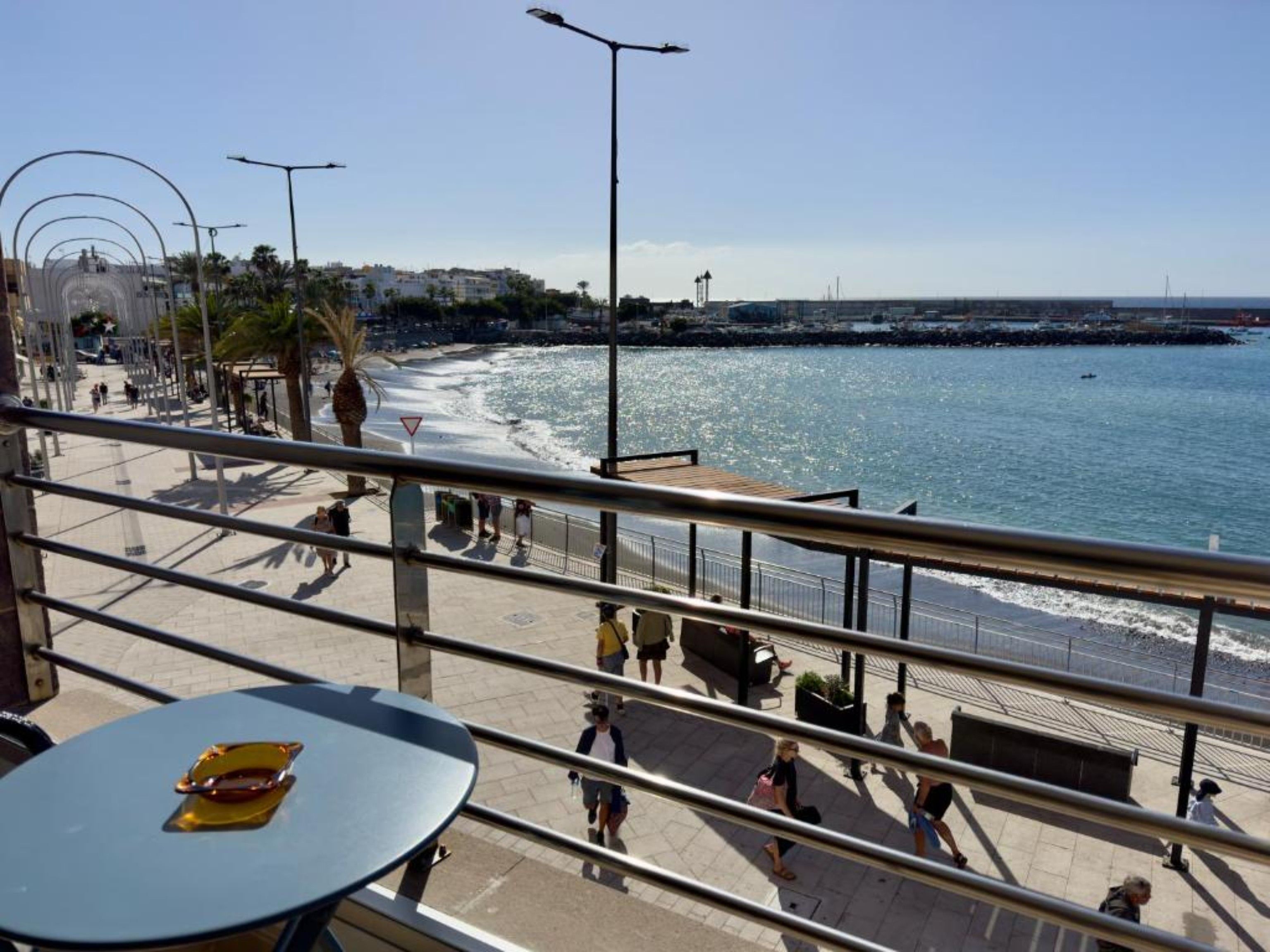 Un paseo marítimo con barandilla, mesas y gente caminando.