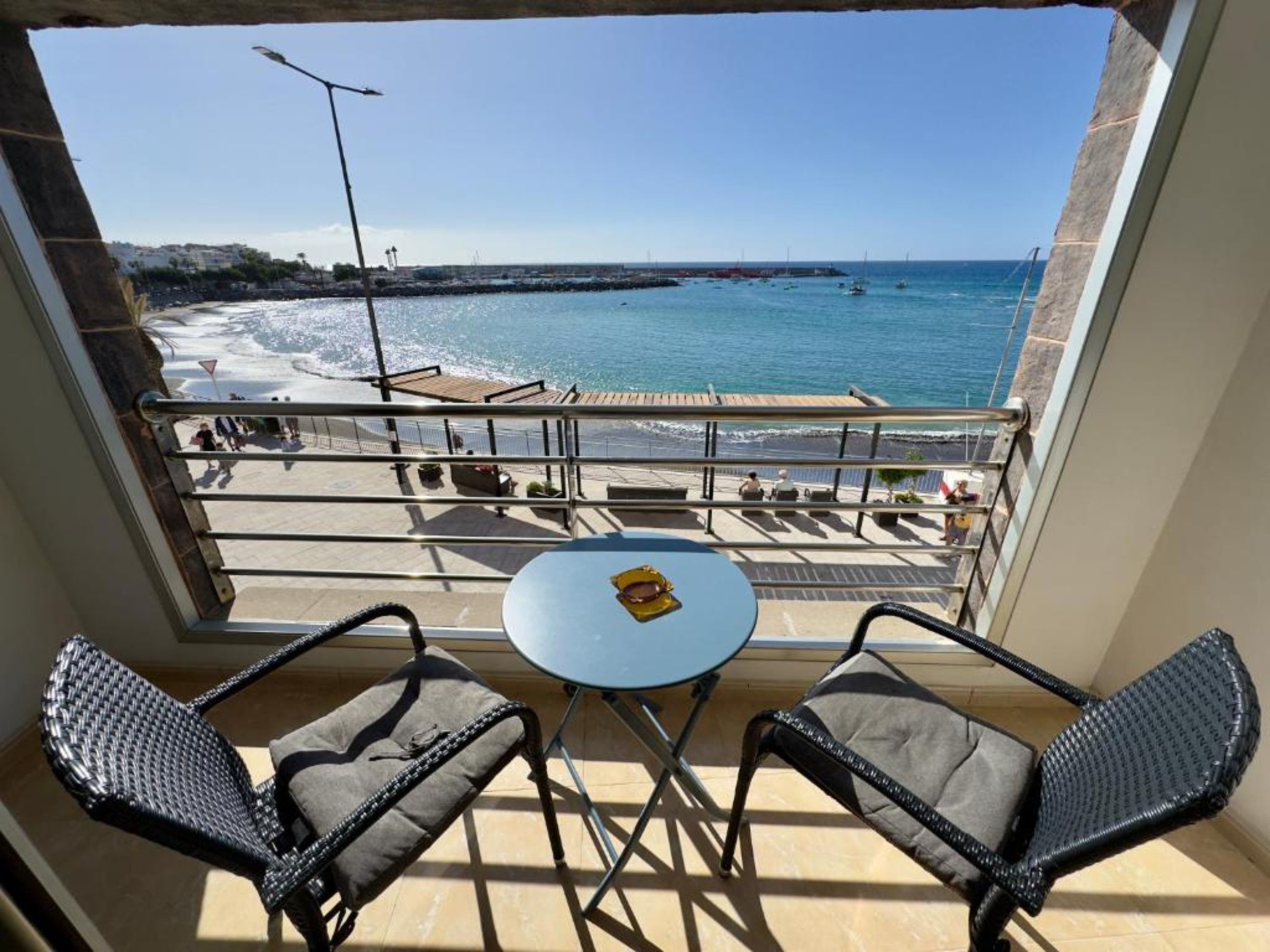 Un balcon avec deux chaises et une petite table, donnant sur une plage où l’on aperçoit des bateaux et un ciel dégagé.
