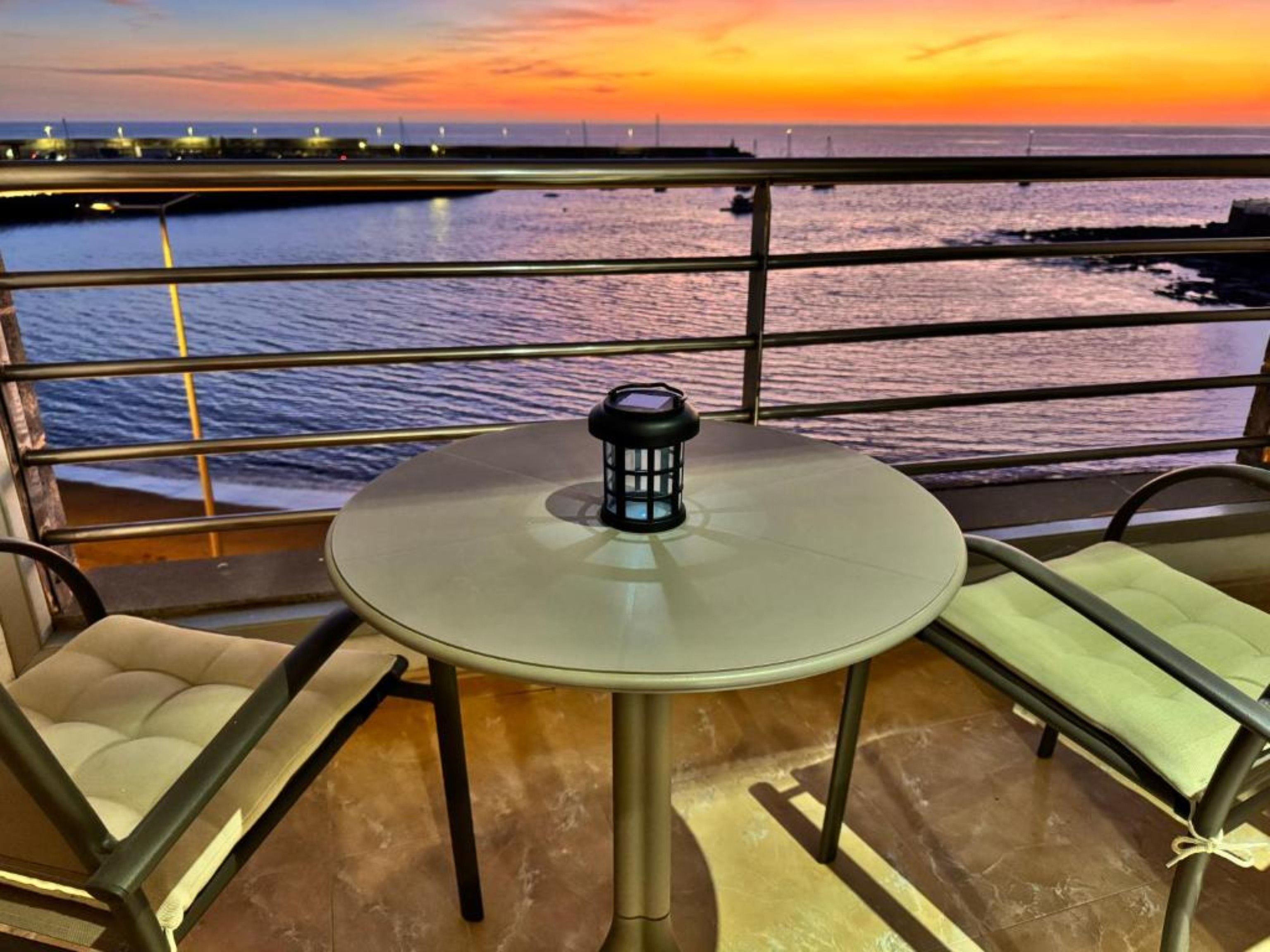 En balkon med et rundt bord og to stole, der vender ud mod vandet i solnedgang.