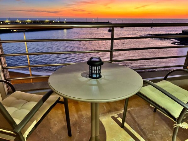 A balcony with a round table and two chairs, overlooking a body of water at sunset.