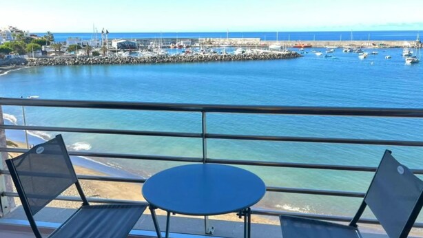 A balcony with two chairs and a small table, overlooking a marina with boats and a rocky breakwater.