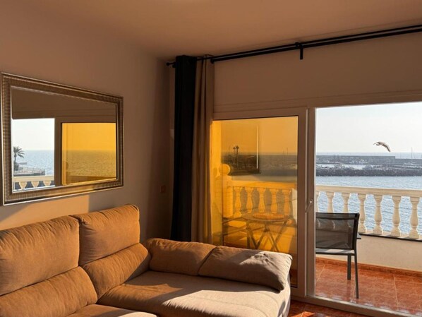 Een kamer met een bank, een stoel, een balkon met hekwerk en uitzicht op zee.