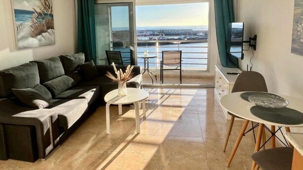 Una sala de estar moderna con sofá, mesa de centro y comedor, con un gran ventanal con vista al mar.