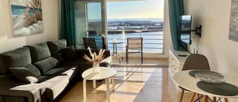 Un salon moderne comprenant un canapé, une table basse et un coin repas, avec une grande fenêtre offrant une vue sur la mer.