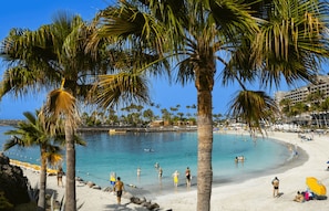 Een strand met palmbomen, helderblauw water en mensen die zwemmen en zonnebaden.