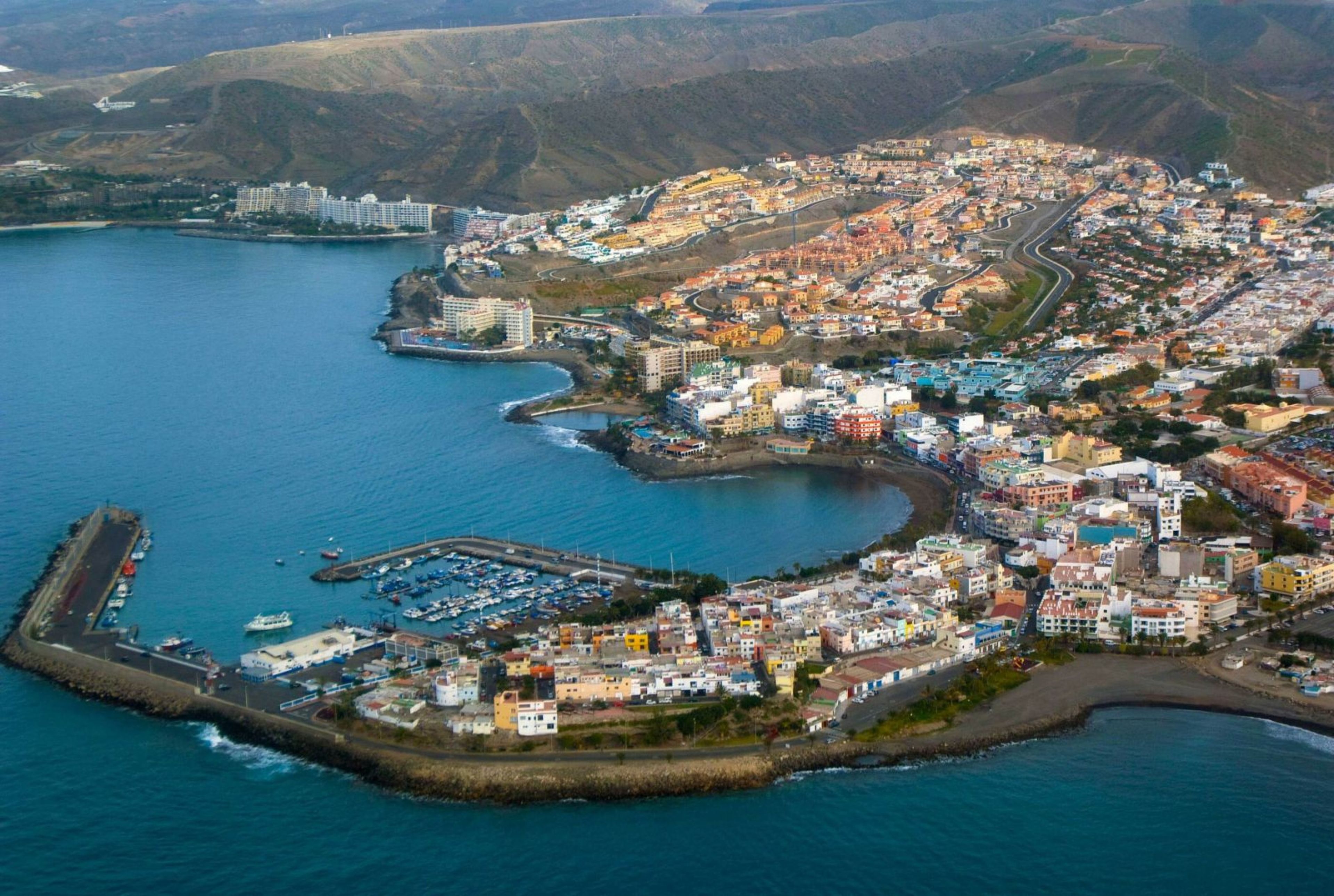 An aerial view of a coastal town with a marina, buildings, and a hilly backdrop.