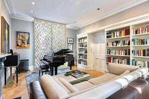 A modern living room with a grand piano, a sofa, a coffee table, and a bookshelf filled with books. - St John s Wood 4-bed House Huge Garden Grand Piano (London)
