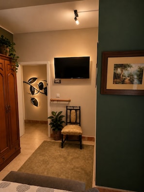 A living room with a flat-screen TV mounted on the wall, a wooden cabinet, a chair, a plant, and a framed picture.